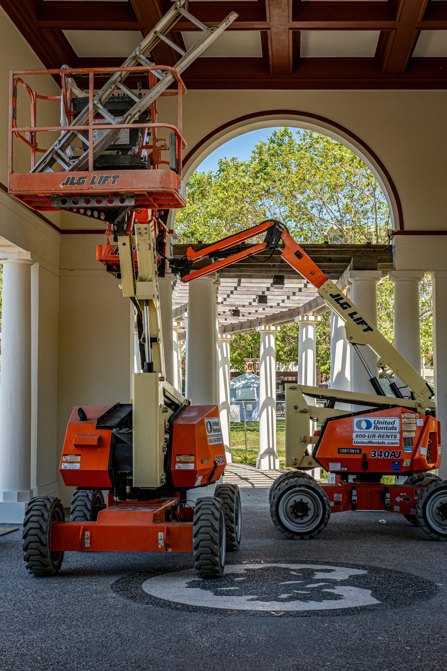 Two orange JLG lifts inside a building with a view outside through an archway.